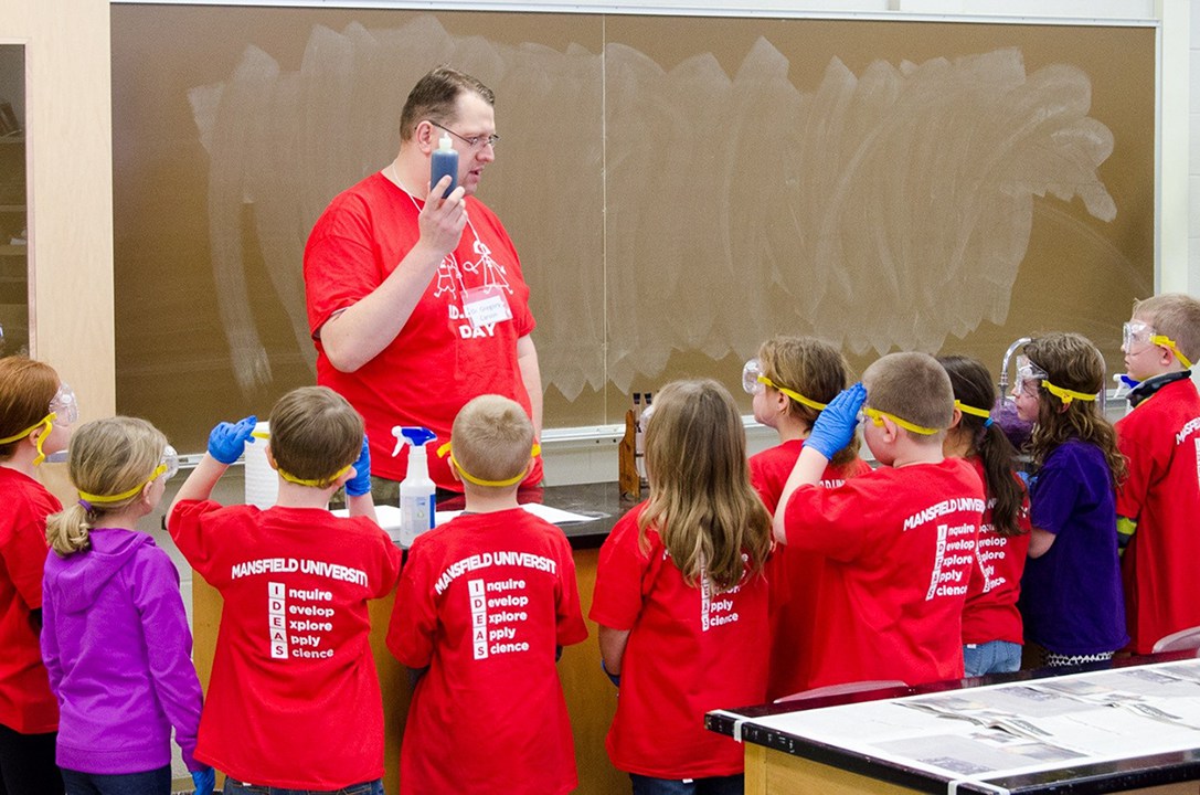 Professor showing a bottle of colored liquid to a group of young students in safety goggles